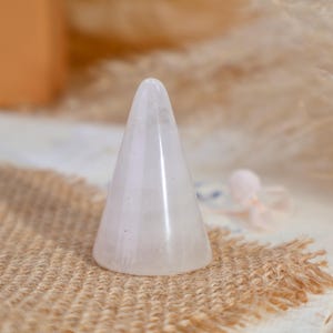 May include: A smooth, conical ring holder crafted from translucent, pale pink rose quartz. The object features a pointed top and a wide, circular base, displaying the natural crystalline texture and soft, milky coloration of the stone material.