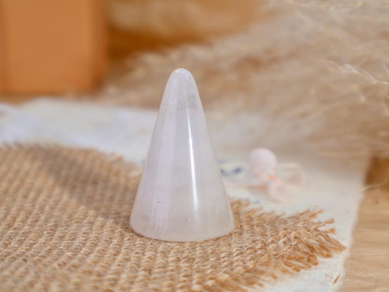 May include: A smooth, conical ring holder crafted from translucent, pale pink rose quartz. The object features a pointed top and a wide, circular base, displaying the natural crystalline texture and soft, milky coloration of the stone material.