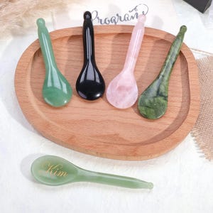 Pode incluir: Cinco colheres de massagem facial em pedra lisa e polida em várias cores, incluindo verde claro, preto, rosa pálido e verde escuro manchado. Uma colher verde claro apresenta o nome Kim gravado em letras douradas na parte côncava.