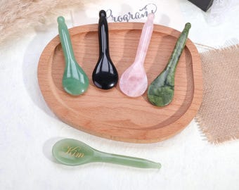 May include: Five smooth, polished stone facial massage spoons in various colors including light green, black, pale pink, and dark mottled green. One light green spoon features the name Kim engraved in gold script on the bowl of the spoon.