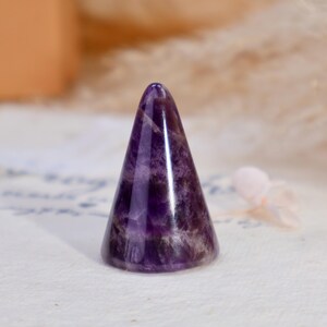 May include: A polished amethyst crystal carved into a smooth, conical shape. The stone features deep purple hues with natural white banding and translucent sections, showcasing a glossy, reflective surface that tapers to a rounded point.