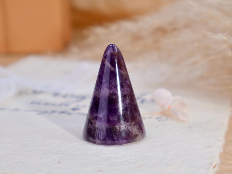 May include: A polished amethyst crystal carved into a smooth, conical shape. The stone features deep purple hues with natural white banding and translucent sections, showcasing a glossy, reflective surface that tapers to a rounded point.