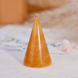 May include: A smooth, polished orange stone ring holder shaped like a cone. The object features a translucent, mottled texture with varying shades of amber and gold throughout its surface, standing upright with a circular base and a pointed top.