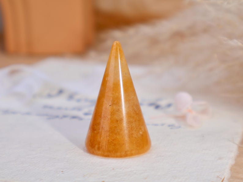 May include: A smooth, polished orange stone ring holder shaped like a cone. The object features a translucent, mottled texture with varying shades of amber and gold throughout its surface, standing upright with a circular base and a pointed top.
