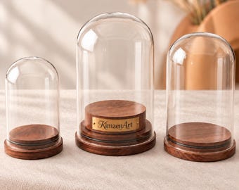 May include: Three clear glass display domes with rounded tops, each resting on a circular dark brown wooden base. The central dome features a small rectangular brass-colored plaque with the engraved text KimzenArt centered on the wooden stand.