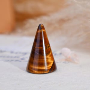 May include: A polished tiger's eye gemstone carved into a smooth, conical shape. The stone features characteristic golden-brown and dark brown banded patterns with a reflective, chatoyant luster that shifts across the surface of the cone.