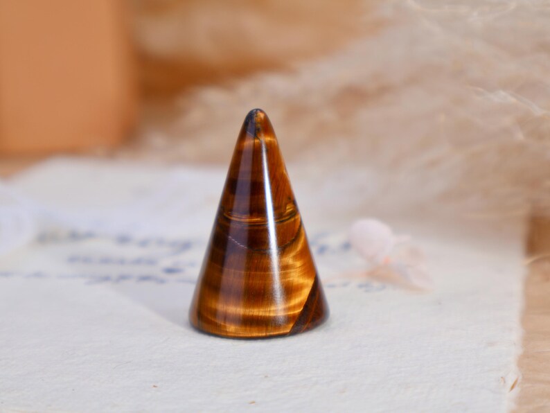 May include: A polished tiger's eye gemstone carved into a smooth, conical shape. The stone features characteristic golden-brown and dark brown banded patterns with a reflective, chatoyant luster that shifts across the surface of the cone.