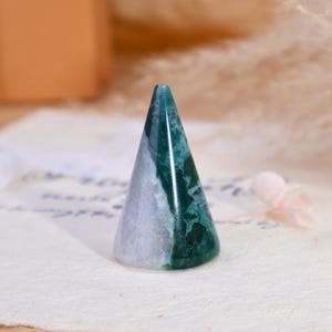May include: A polished stone ring holder in a conical shape. The object features a distinct color split, with one side displaying a translucent light lavender hue and the other side showcasing a deep forest green with intricate moss-like inclusions.