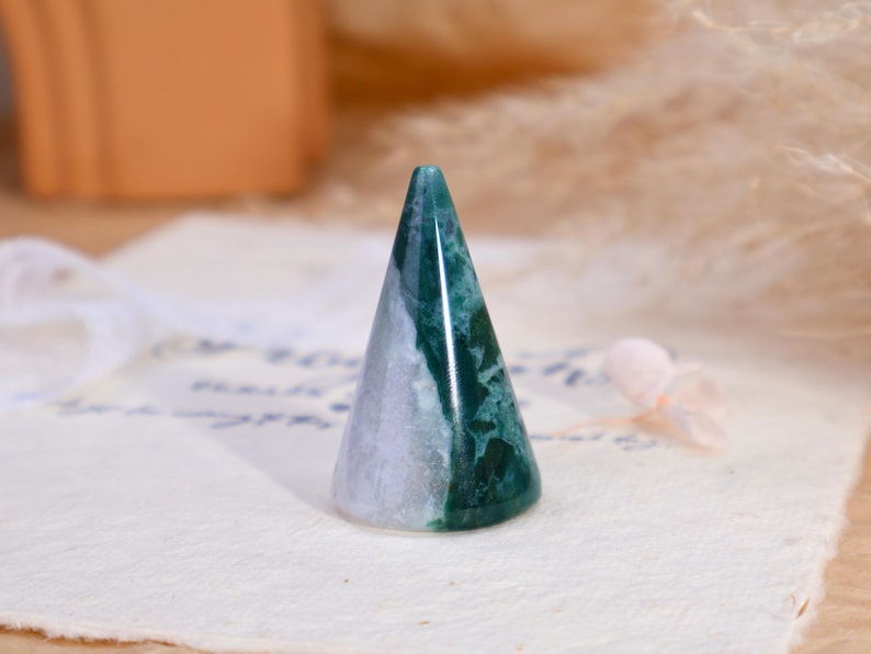 May include: A polished stone ring holder in a conical shape. The object features a distinct color split, with one side displaying a translucent light lavender hue and the other side showcasing a deep forest green with intricate moss-like inclusions.