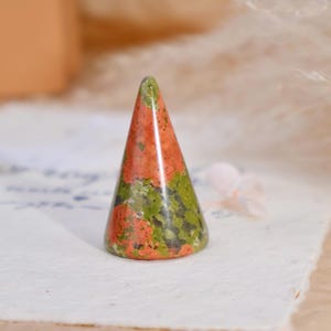 May include: A cone-shaped ring holder carved from unakite stone, featuring a mottled pattern of earthy green and salmon pink colors with a smooth, polished finish. The object stands upright with a circular base and tapers to a rounded point.