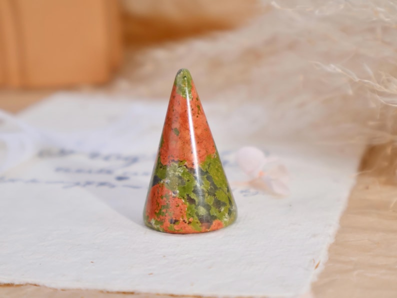 May include: A cone-shaped ring holder carved from unakite stone, featuring a mottled pattern of earthy green and salmon pink colors with a smooth, polished finish. The object stands upright with a circular base and tapers to a rounded point.