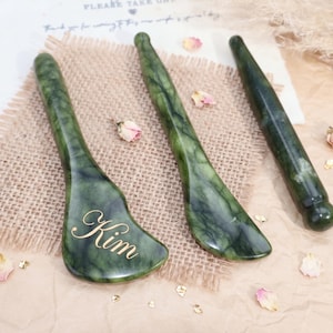 Peut inclure: Trois outils de massage en pierre polie vert foncé avec une texture marbrée. Un outil est large et plat avec le nom Kim gravé en écriture dorée, tandis que les deux autres sont des baguettes cylindriques longues aux extrémités arrondies.