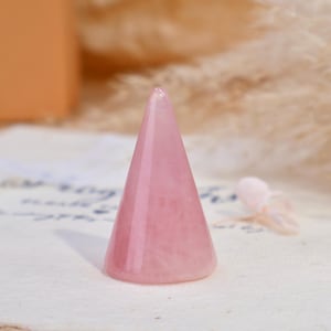May include: A smooth, polished rose quartz crystal carved into a conical shape. The stone features a translucent, pale pink hue with natural internal variations and a pointed top, designed as a stand for displaying rings or other small jewelry items.