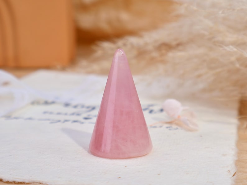 May include: A smooth, polished rose quartz crystal carved into a conical shape. The stone features a translucent, pale pink hue with natural internal variations and a pointed top, designed as a stand for displaying rings or other small jewelry items.