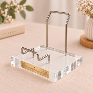 May include: A clear acrylic display stand featuring a silver-toned metal frame for holding items. The base includes a rectangular gold-colored plaque with the text Moonlight Crystal and decorative leaf illustrations on each side.