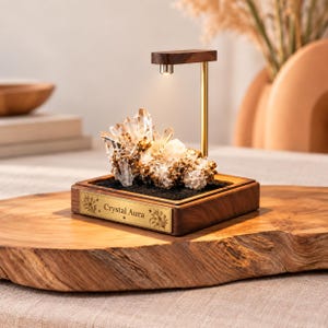 May include: A clear quartz crystal cluster sits on a dark base within a wooden frame. A gold-toned metal arm extends upward with a small light fixture pointing down. A gold plaque on the front of the wooden base features the text Crystal Aura.