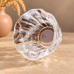 May include: A clear glass bowl featuring a textured, irregular surface with a rippled, organic rim. The glass has a thick, heavy base and a translucent appearance, catching light to highlight the uneven, crystalline-like patterns on its exterior walls.