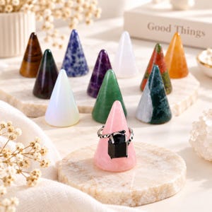 May include: A collection of cone-shaped ring holders crafted from various polished stones, including rose quartz, amethyst, sodalite, and tiger's eye. A silver-toned ring featuring a large, rectangular black stone is displayed on the pink cone.