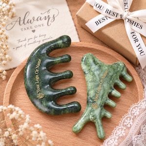Peut inclure: Deux outils de massage en pierre verte polie avec plusieurs dents. Chaque outil porte le texte doré Your Logo Or Name et une petite icône de fleur de lotus. Les outils sont lisses, incurvés et ont une finition brillante sur un plateau en bois.