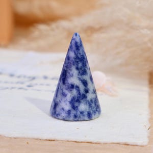 May include: A cone-shaped ring holder crafted from polished stone, featuring a mottled pattern of deep blue and white colors. The object has a smooth, tapered surface and a circular base, designed to hold rings securely on its pointed top.