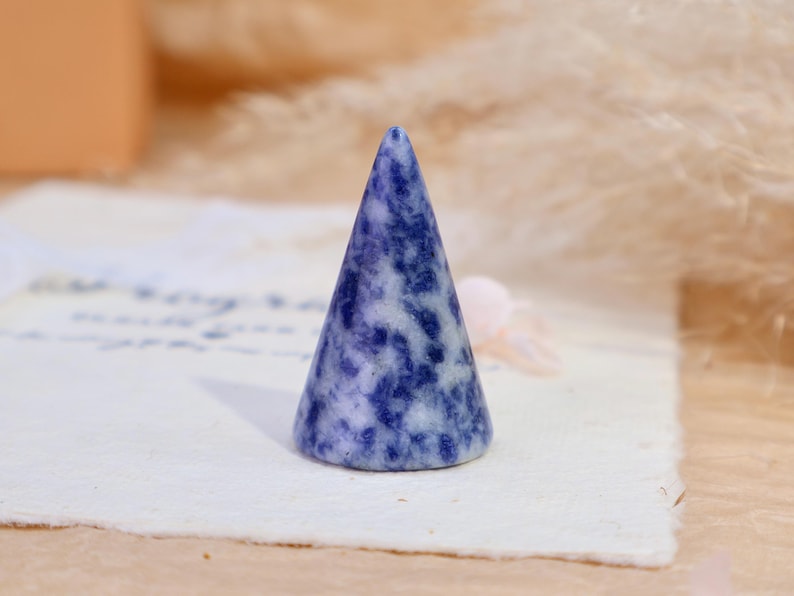 May include: A cone-shaped ring holder crafted from polished stone, featuring a mottled pattern of deep blue and white colors. The object has a smooth, tapered surface and a circular base, designed to hold rings securely on its pointed top.