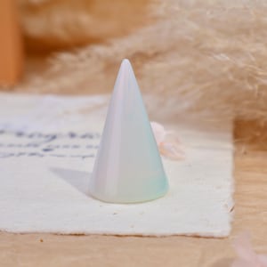 May include: A smooth, conical ring display stand with an iridescent, pearlescent finish that shifts between soft white, pale blue, and subtle green tones. The object has a wide, circular base that tapers to a rounded point at the top.