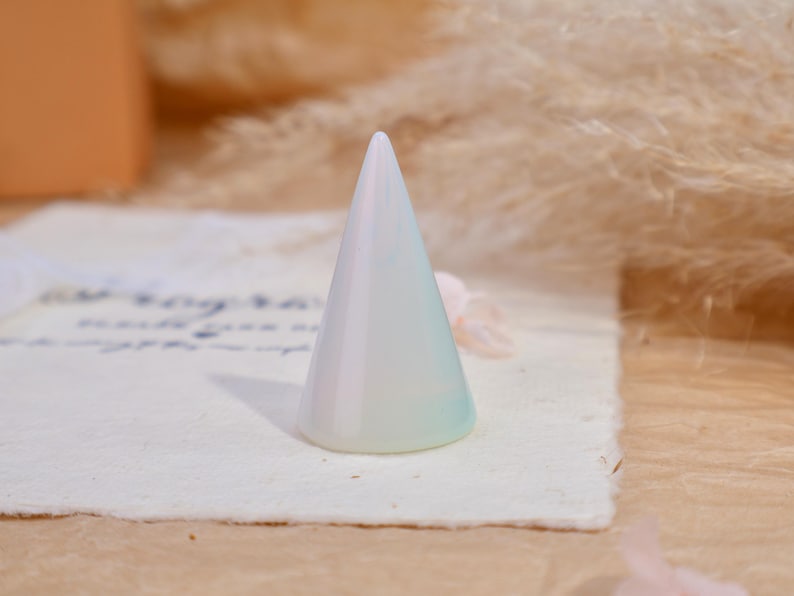 May include: A smooth, conical ring display stand with an iridescent, pearlescent finish that shifts between soft white, pale blue, and subtle green tones. The object has a wide, circular base that tapers to a rounded point at the top.