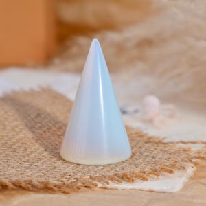 May include: A smooth, conical ring holder made of translucent, milky white material with a soft iridescent sheen. The object features a wide, circular base that tapers to a rounded point at the top, designed for displaying rings in an upright position.