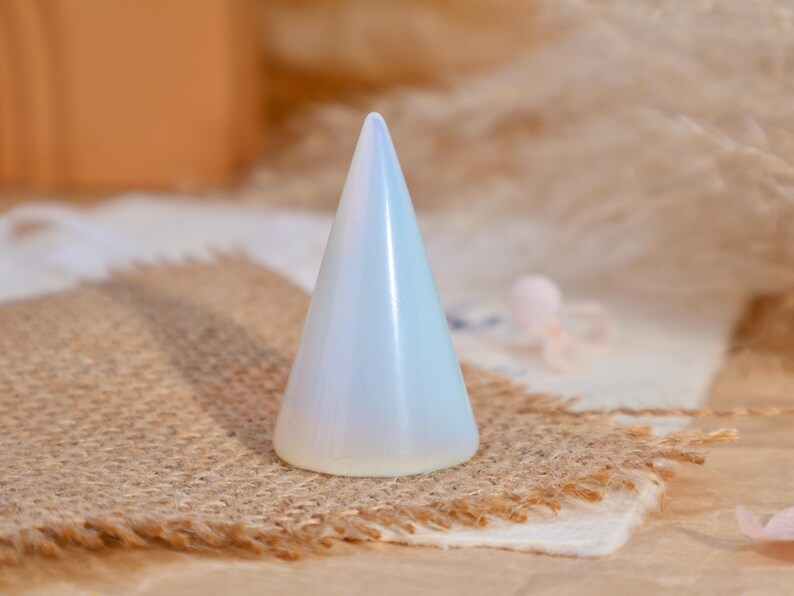 May include: A smooth, conical ring holder made of translucent, milky white material with a soft iridescent sheen. The object features a wide, circular base that tapers to a rounded point at the top, designed for displaying rings in an upright position.