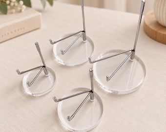 May include: A set of four display stands, each featuring a clear, circular acrylic base and a silver-toned metal support structure. The stands vary in size, with each unit consisting of a vertical rod and two angled arms designed to hold items upright.