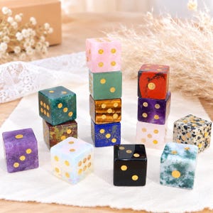 May include: A collection of twelve multi-colored stone dice featuring gold-colored pips. The dice are crafted from various minerals including pink, green, brown, blue, purple, black, and speckled white stones, arranged in stacks and individually.