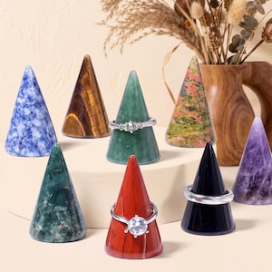 May include: A collection of eight polished stone ring cones in various colors including blue, brown, green, red, black, and purple. Three of the cones are adorned with silver-toned rings, featuring clear stones and intricate metal band designs.