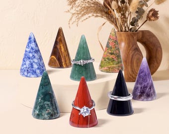 Op de afbeelding: Een verzameling van acht gepolijste stenen ringkegels in diverse kleuren, waaronder blauw, bruin, groen, rood, zwart en paars. Drie van de kegels zijn versierd met zilverkleurige ringen, voorzien van heldere stenen en gedetailleerde metalen ontwerpen.