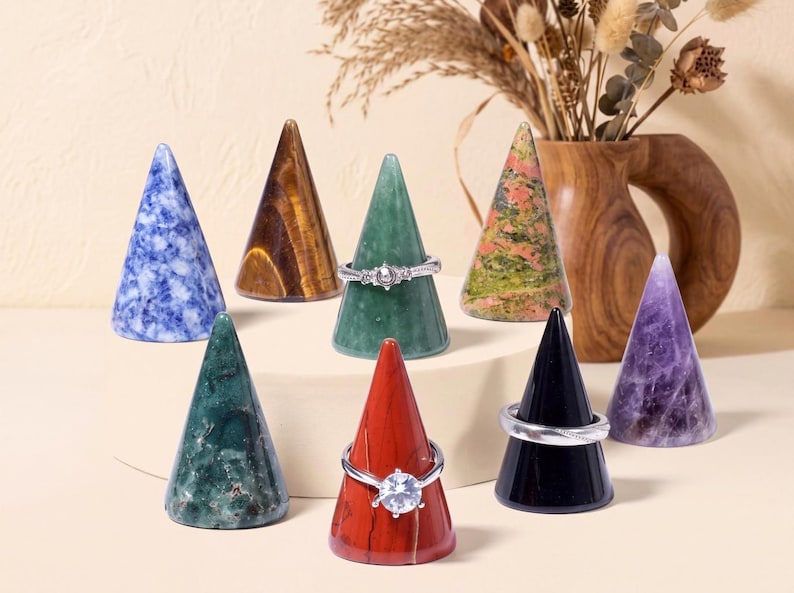 May include: A collection of eight polished stone ring cones in various colors including blue, brown, green, red, black, and purple. Three of the cones are adorned with silver-toned rings, featuring clear stones and intricate metal band designs.