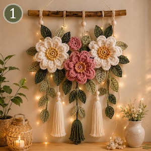 May include: A wall hanging featuring crocheted flowers in white and pink with yellow centers, surrounded by dark green crocheted leaves. The arrangement is suspended from a wooden branch with light-colored wooden beads and three cream-colored tassels.