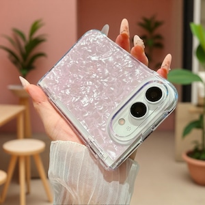 Op de afbeelding: Een opvouwbaar smartphonehoesje met een lichtroze, iriserende textuur van vermalen schelpen. Het hoesje heeft een transparant beschermend frame en een uitsparing voor het dubbele camerasysteem en de flitser aan de achterzijde.