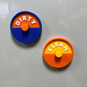 May include: Two circular dishwasher magnets with a rotating center knob. One magnet has a blue outer ring, an orange top half with the white text DIRTY, and a blue bottom half. The other has an orange outer ring, a yellow top half with the white text CLEAN, and an orange bottom half.
