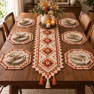 Puede incluir: Un camino de mesa de ganchillo y seis manteles individuales octogonales a juego en tonos crema, naranja y óxido. El camino presenta un motivo floral repetitivo con borlas decorativas en los extremos. Cada mantel sostiene un plato blanco.