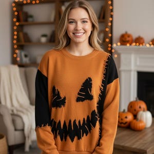 May include: A long-sleeve crewneck jumper featuring a pumpkin jack-o'-lantern face design. The jumper is primarily orange with black sleeves and black jagged eye and mouth details. It includes visible stitched seam accents along the front.