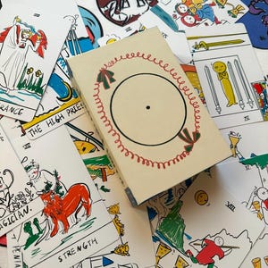 Puede incluir: Una baraja de cartas de tarot con ilustraciones dibujadas a mano en una paleta de colores blanco, rojo, verde y amarillo. La caja central es de color crema con un diseño de corona roja circular. Las cartas muestran figuras como Strength, The High Priestess y The Magician.