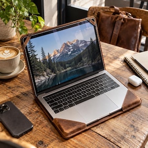 May include: A silver laptop secured in a brown leather protective case with corner straps. The screen displays a landscape image of a mountain range with snow-capped peaks, a dense forest of pine trees, and a calm lake reflecting the scenery.