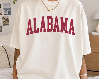 May include: An off-white short-sleeve t-shirt featuring the word ALABAMA printed in a large, arched, dark red collegiate-style font across the chest. The shirt has a crew neckline and a relaxed, loose fit, paired with a gold-toned chain necklace.