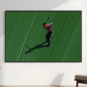 Puede incluir: Una obra de arte mural enmarcada que presenta una pintura al óleo de un golfista con camisa roja y pantalones negros balanceando un palo de golf. La figura se sitúa sobre un fondo verde intenso texturizado con una larga sombra oscura.