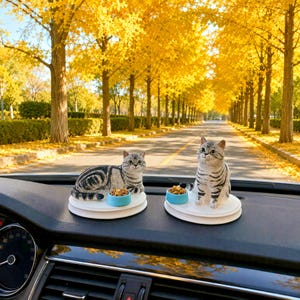 May include: Two grey and black tabby cat figurines displayed on white circular bases. Each figurine is accompanied by a small, light blue bowl filled with brown kibble. The cats are positioned on a dark dashboard inside a vehicle.
