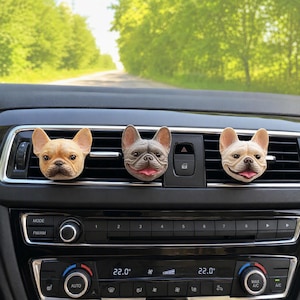 May include: Three French bulldog head car air vent clips attached to a vehicle dashboard. The left clip is a tan dog, the centre is a grey dog with its tongue out, and the right is a white dog with its tongue out, all featuring detailed faces.