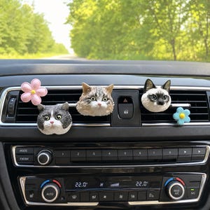 May include: Three cat head air freshener clips and two flower clips attached to a car dashboard air vent. The cat heads include a grey and white tabby, a brown and white tabby, and a Siamese cat. The flowers are light pink and light blue.