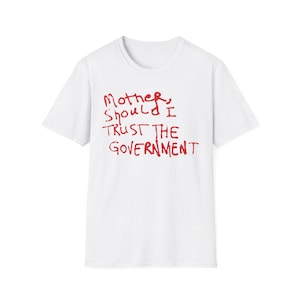 May include: White t-shirt with the red text "Mother, Should I Trust The Government" in a handwritten style. The shirt has a crew neck and short sleeves. The text is centered on the front of the shirt.