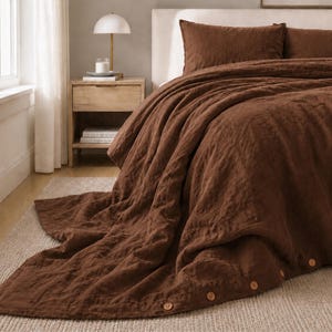 May include: A dark brown duvet cover featuring a textured fabric surface and a row of small, round, light-colored buttons along the bottom edge. The bedding set includes matching pillowcases, all displayed on a bed with a neutral-colored headboard.