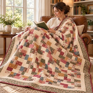 May include: A patchwork quilt featuring a repeating pattern of small floral squares in shades of red, blue, yellow, and green, bordered by a dark green floral trim. The quilt is draped over a person reading a book, showcasing intricate stitching.