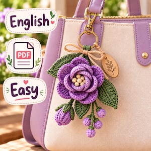 May include: A crochet flower keychain featuring a large purple bloom with a cream-colored center, green leaves, a purple bud, and small purple berries. It includes a beige bow, a wooden oval tag with a leaf engraving, and a gold-tone metal clasp.